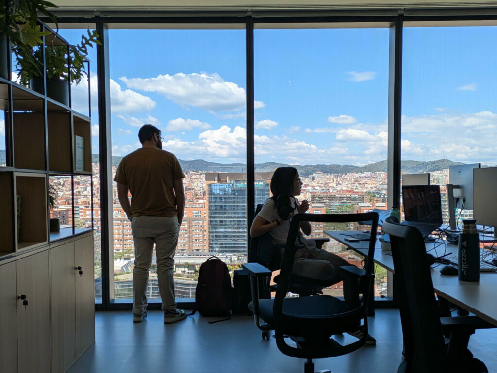View of interior of an office. Window to the outside in the background where we can see a view of the city. The inside is rather dark. Two human silhouette detach from the clear window. One is standing and looking outside. The other is sitting in front of a deck.