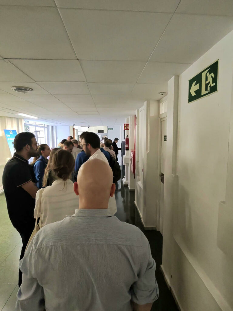 View from a queue with lots of people seen from the back of the queue. Far away, we can see a person turned right and interacting with a machine.